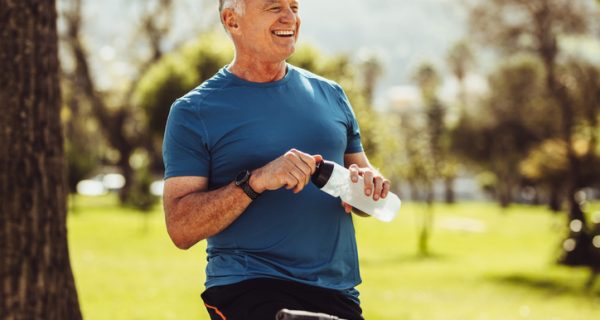 Senior man in fitness wear drinking water sitting on his bicycle. Cheerful senior fitness person taking a break during cycling in a park.